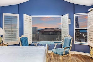 Ein Schlafzimmer mit einem Bett, zwei Stühlen, einem kleinen Tisch und einem großen Fenster mit Rollläden.