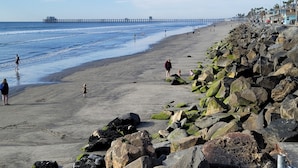 Une plage au rivage rocheux, une jetée en arrière-plan et des personnes se promenant le long du rivage.