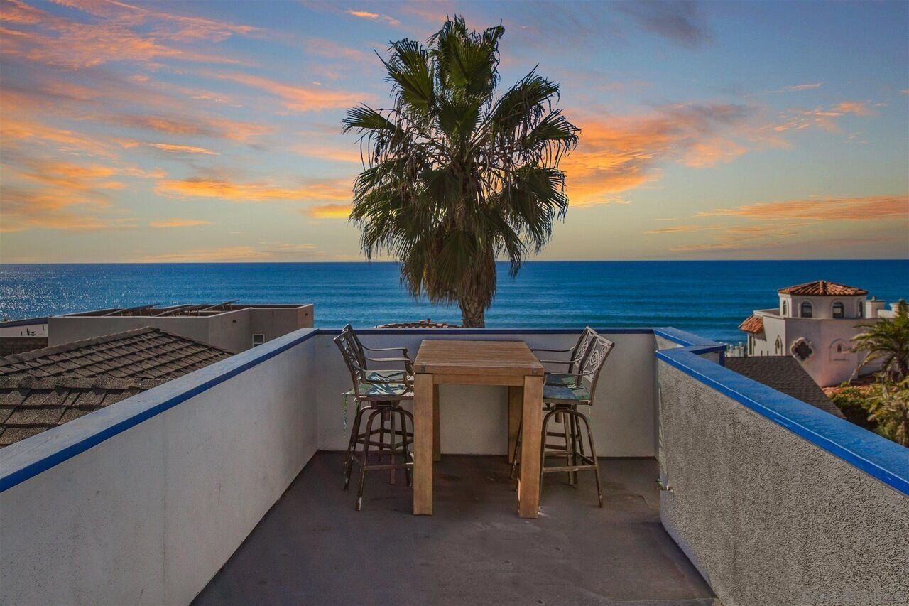 A balcony with a wooden table and chairs, overlooking a palm tree and the sea during sunset.