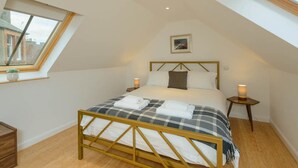 A bedroom with a sloped ceiling, a bed with a wooden frame, two bedside tables with lamps, a window with a view of buildings, and a small plant on the windowsill.