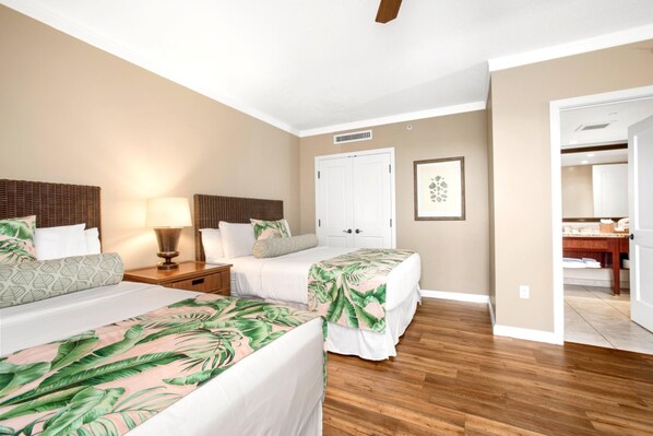 Habitación de hotel con dos camas, cabecera de madera, mesita de noche, lámpara, puerta y baño visible a través de una puerta abierta.
