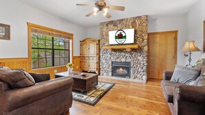 A cozy living room with a stone fireplace, wooden furniture, and a large window. - Newly Remodeled! Cedar Log Cabin with Gas Fireplace & Screened Porch (Branson)