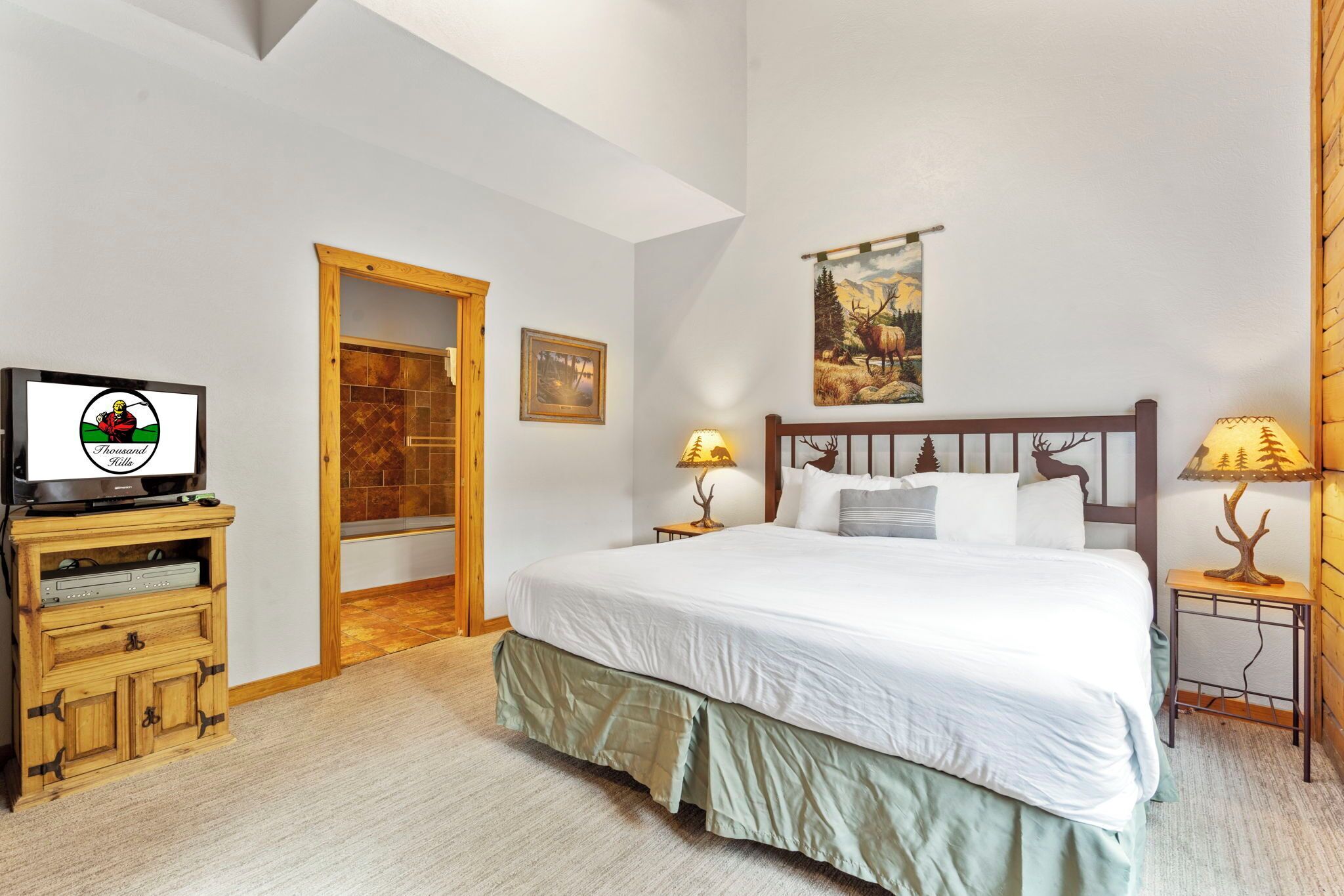 A bedroom with a bed, two bedside lamps, a television on a wooden cabinet, and a door leading to another room.