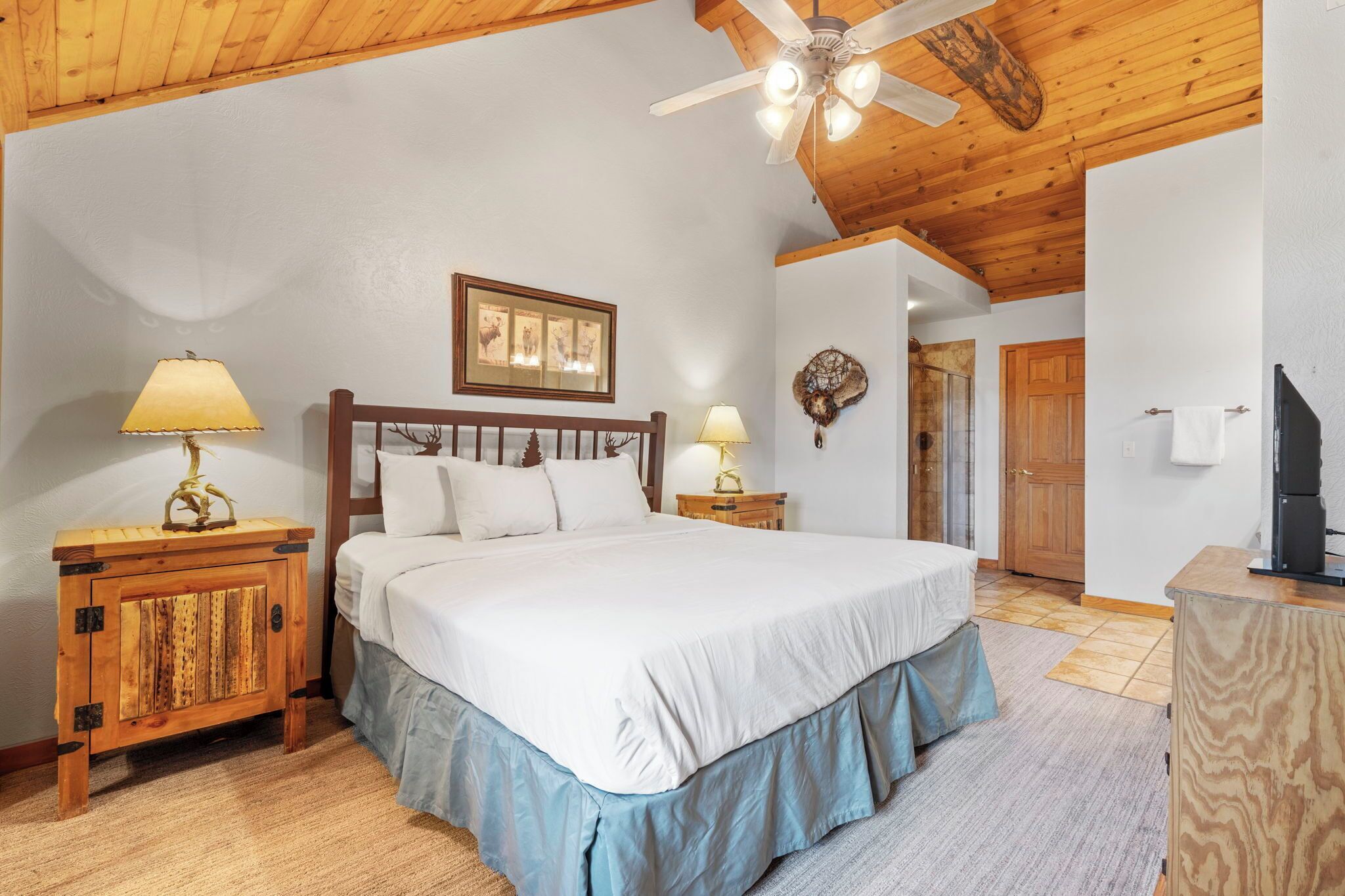 A bedroom with a wooden ceiling, a bed with white bedding, two bedside tables with lamps, and a wooden cabinet.