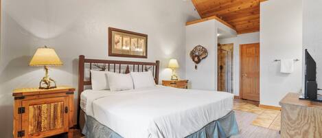 A bedroom with a wooden ceiling, a bed with white bedding, two bedside tables with lamps, and a wooden cabinet.