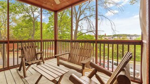 A covered porch with wooden Adirondack chairs and a table, overlooking a street with trees and a building in the background. - Newly Remodeled! Cedar Log Cabin with Gas Fireplace & Screened Porch (Branson)