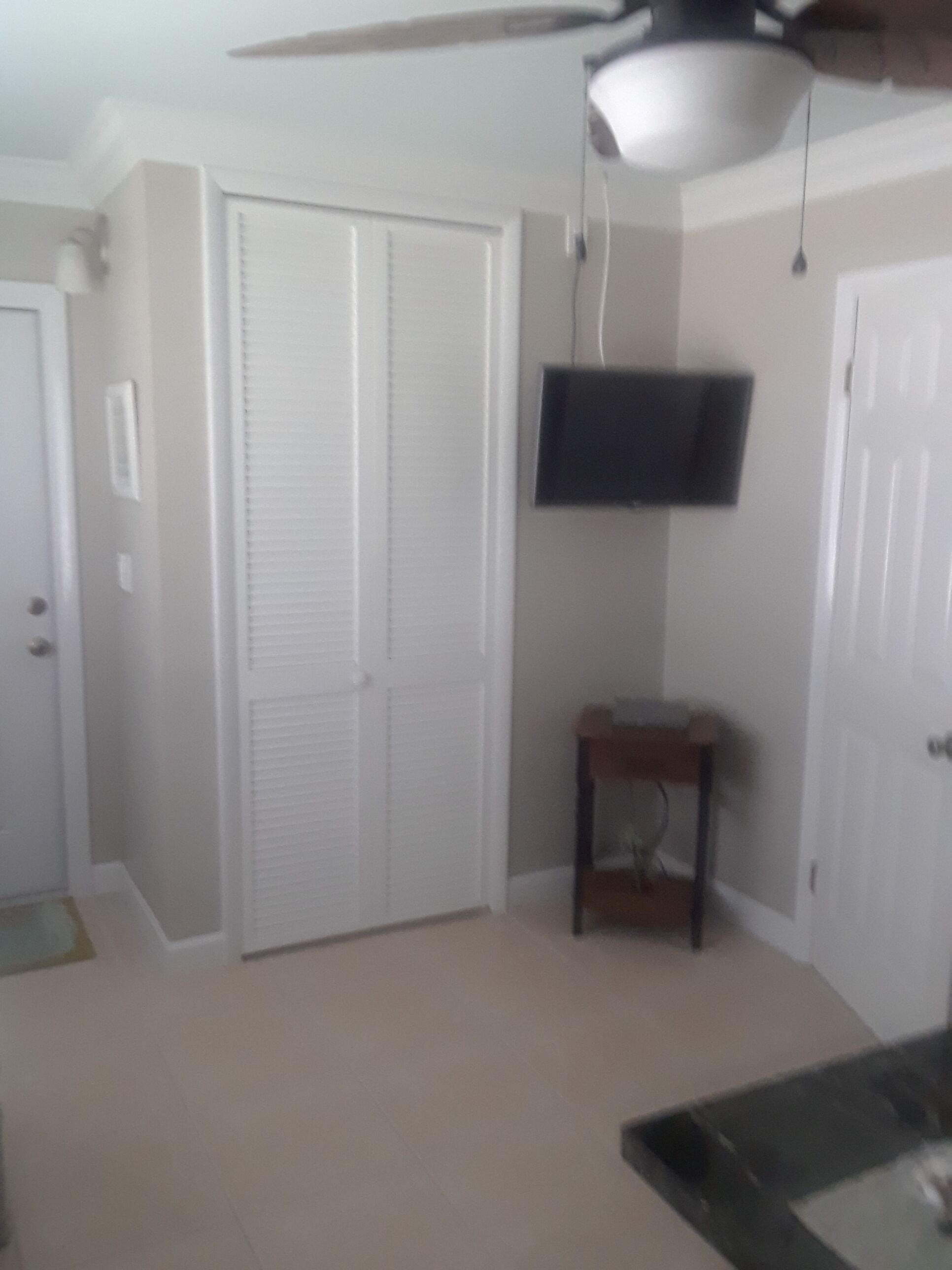 A room with a ceiling fan, a television mounted on the wall, a closed closet, and a small table.