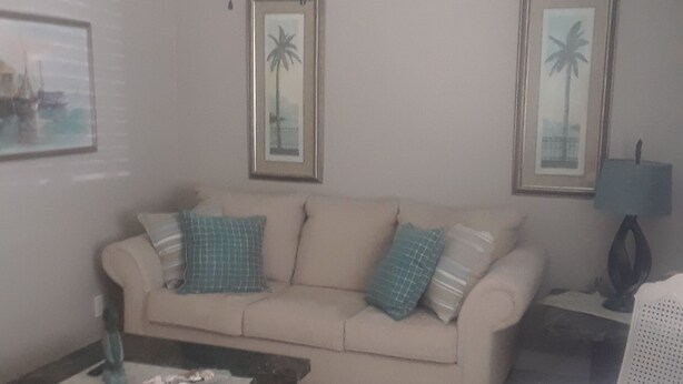 A living room with a beige sofa, a coffee table, and a ceiling fan. There are two framed pictures on the wall.