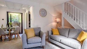 A living room with a grey sofa and armchair, a dining table with chairs, a clock on the wall, and a staircase. - A stylish getaway for discerning guests looking for a great coastal break. (Cromer)