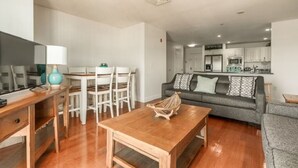 A modern living room with a wooden coffee table, white dining table, and chairs. A grey sofa and a TV are also visible.