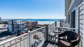 A balcony with a view of the beach and ocean. There are buildings in the background, and a chair is visible on the balcony.