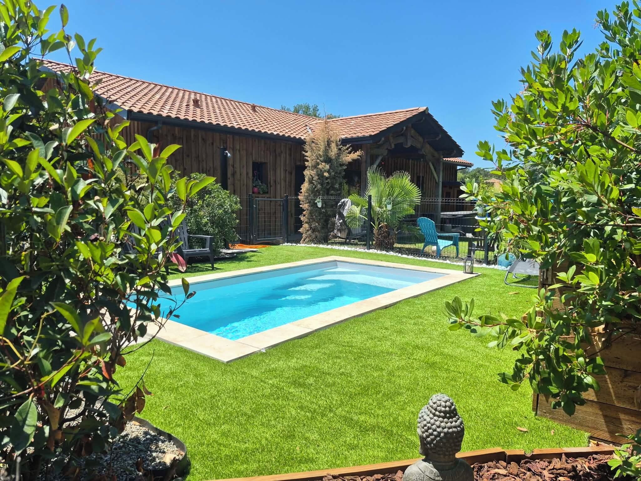 A swimming pool surrounded by artificial grass, a wooden building with a tiled roof, and various plants.