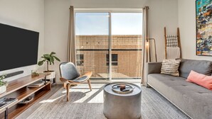 A modern living room with a large flat-screen TV, a grey sofa, a round coffee table, and a large window with a view of a brick building. - LCF - Downtown SLC - 1BR/1BA - CoWork + Gym - Near Delta Center - Garage (Salt Lake City)