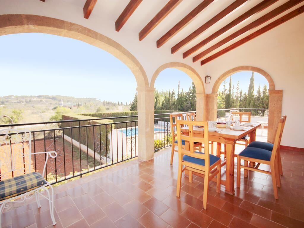 A balcony with a wooden table and chairs, a view of the countryside, and a pool in the background.