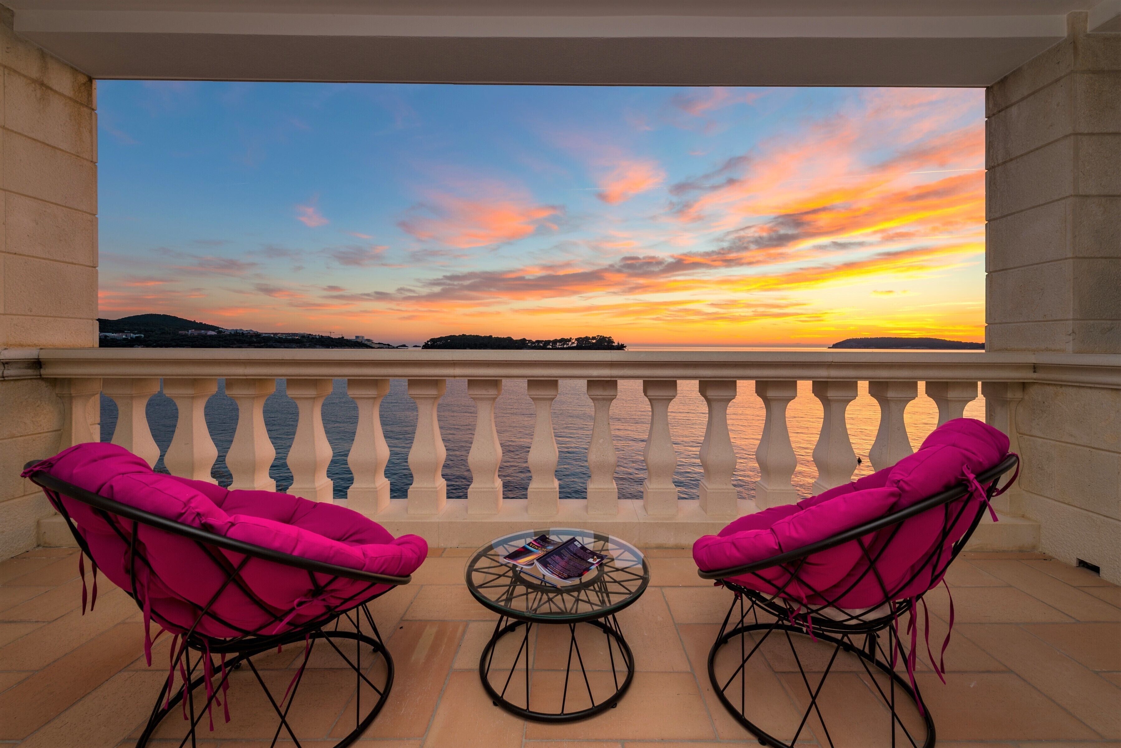 A balcony with two rocking chairs and a small table, overlooking a sunset over the sea.