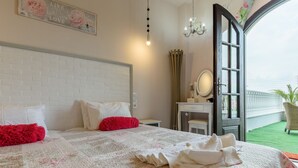 Une chambre d’hôtel avec une tête de lit en briques blanches, un grand lit recouvert d’une literie à motifs floraux blancs et roses, des oreillers rouges et un lit parfaitement fait avec des serviettes. On y trouve une commode avec un miroir, un lustre et, par une porte ouverte, une vue sur une pelouse verdoyante et des palmiers.