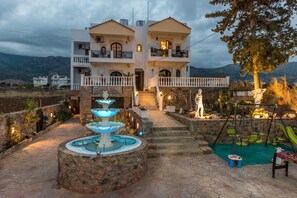 A two-story building with a stone pathway leading to a fountain, a swing set, and a statue. - Olympus Studios Afrodite (Hersonissos)