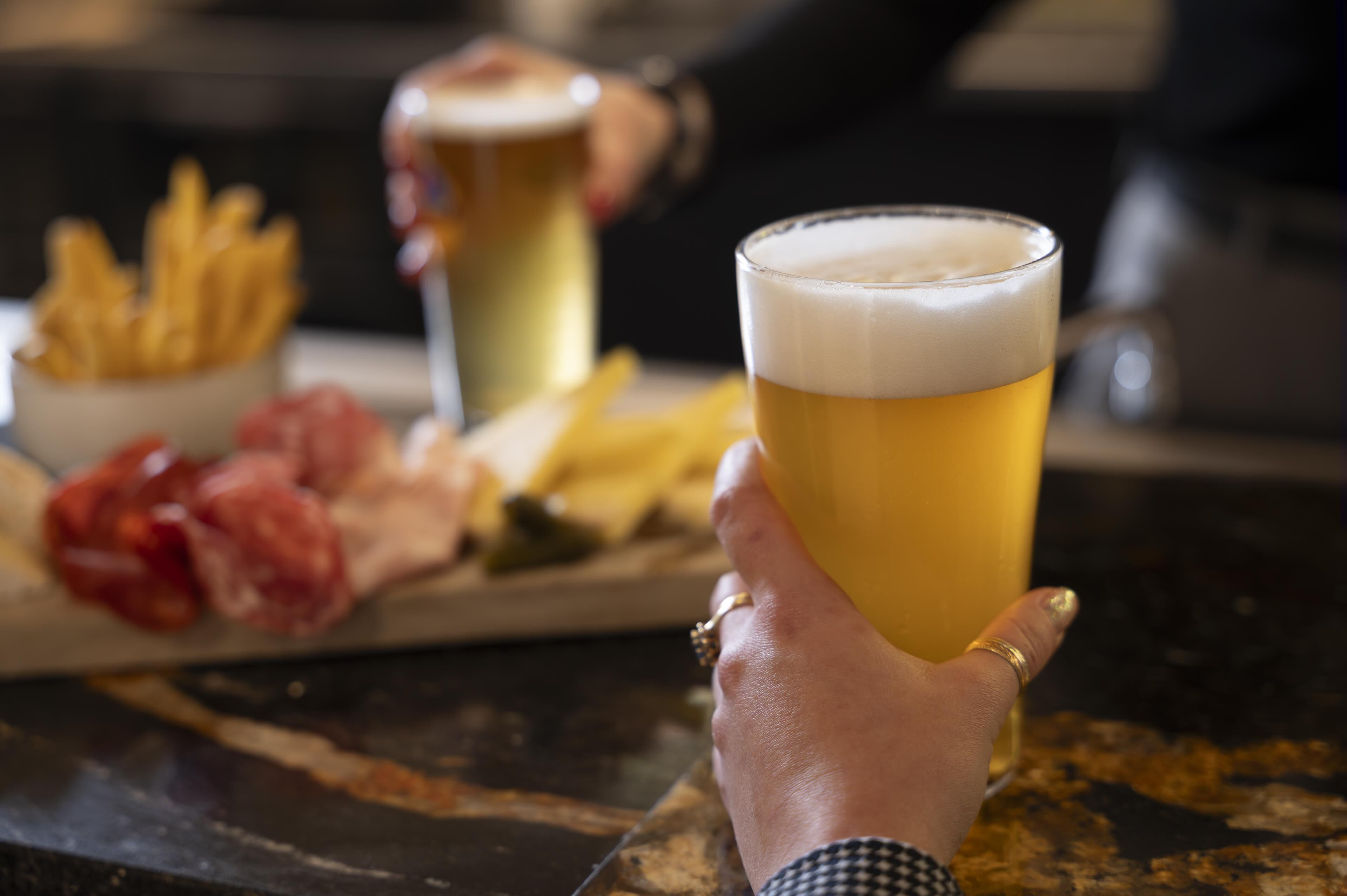 Una mano che tiene un bicchiere di birra, con un piatto di cibo sullo sfondo.