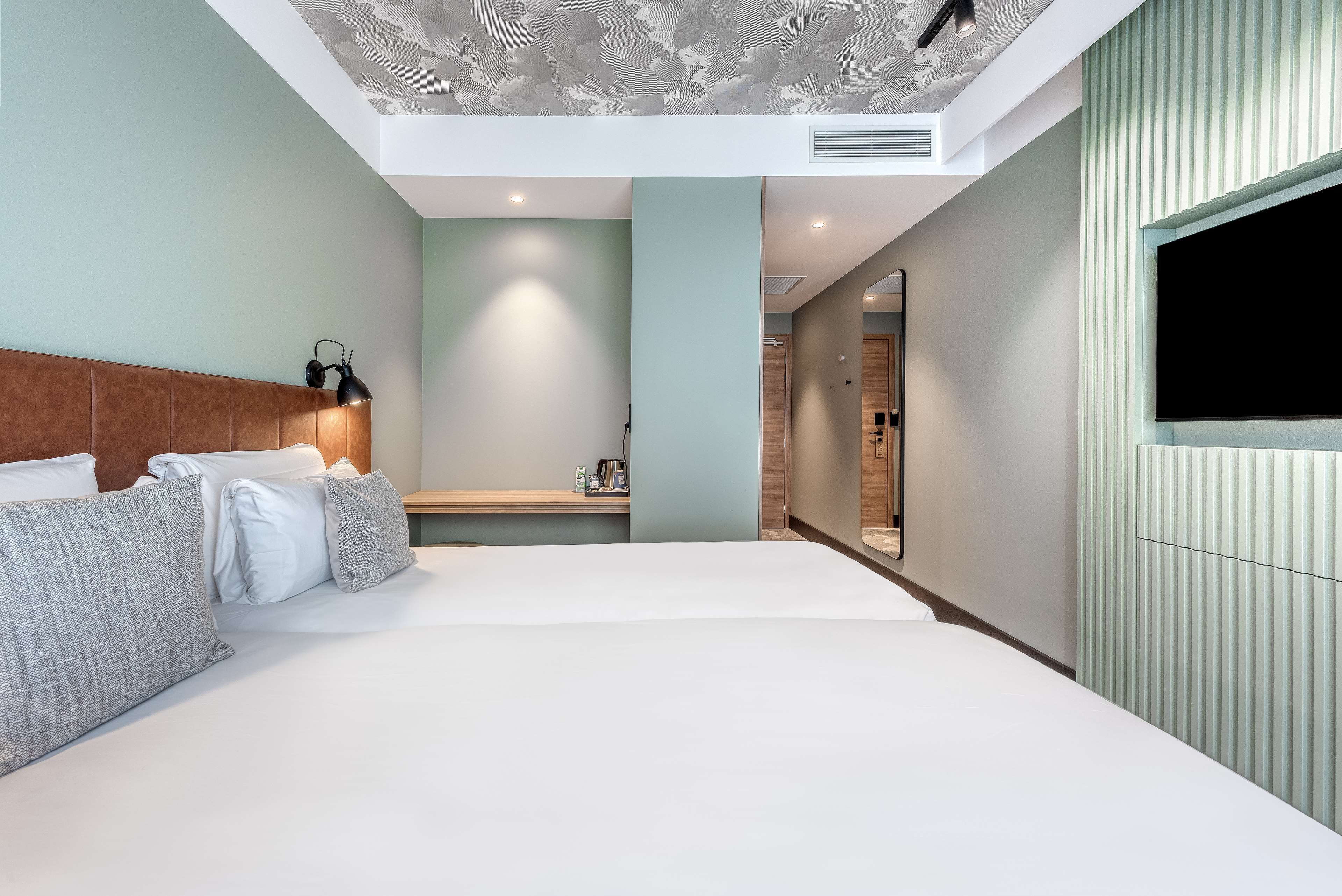 Una habitación de hotel moderna con una cama grande, un cabecero, una mesita de noche, un televisor y un baño visible a través de una puerta abierta.