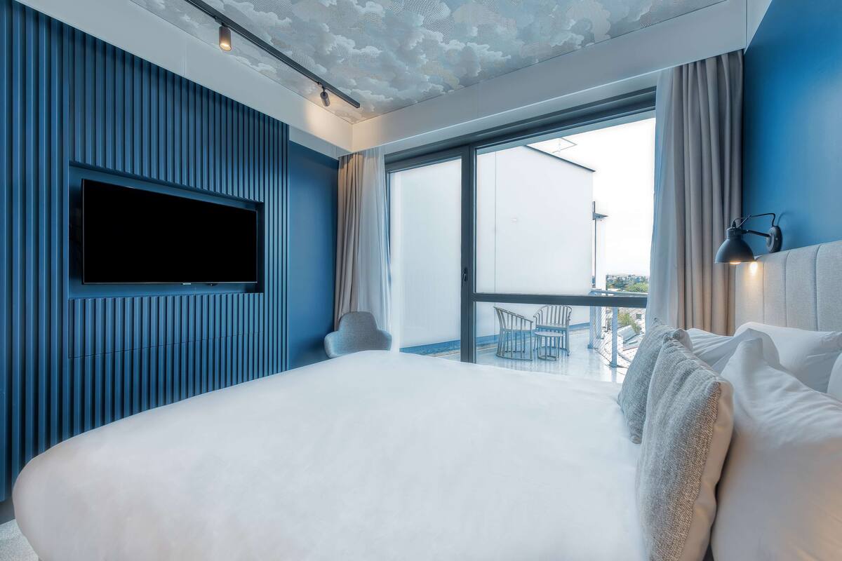 A modern hotel room with a large bed, a flat-screen TV mounted on the wall, and a balcony with a view of the city.