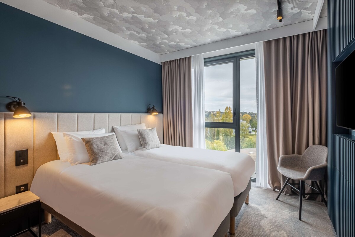 A hotel room with two beds, a view of the outdoors, and a chair by the window.