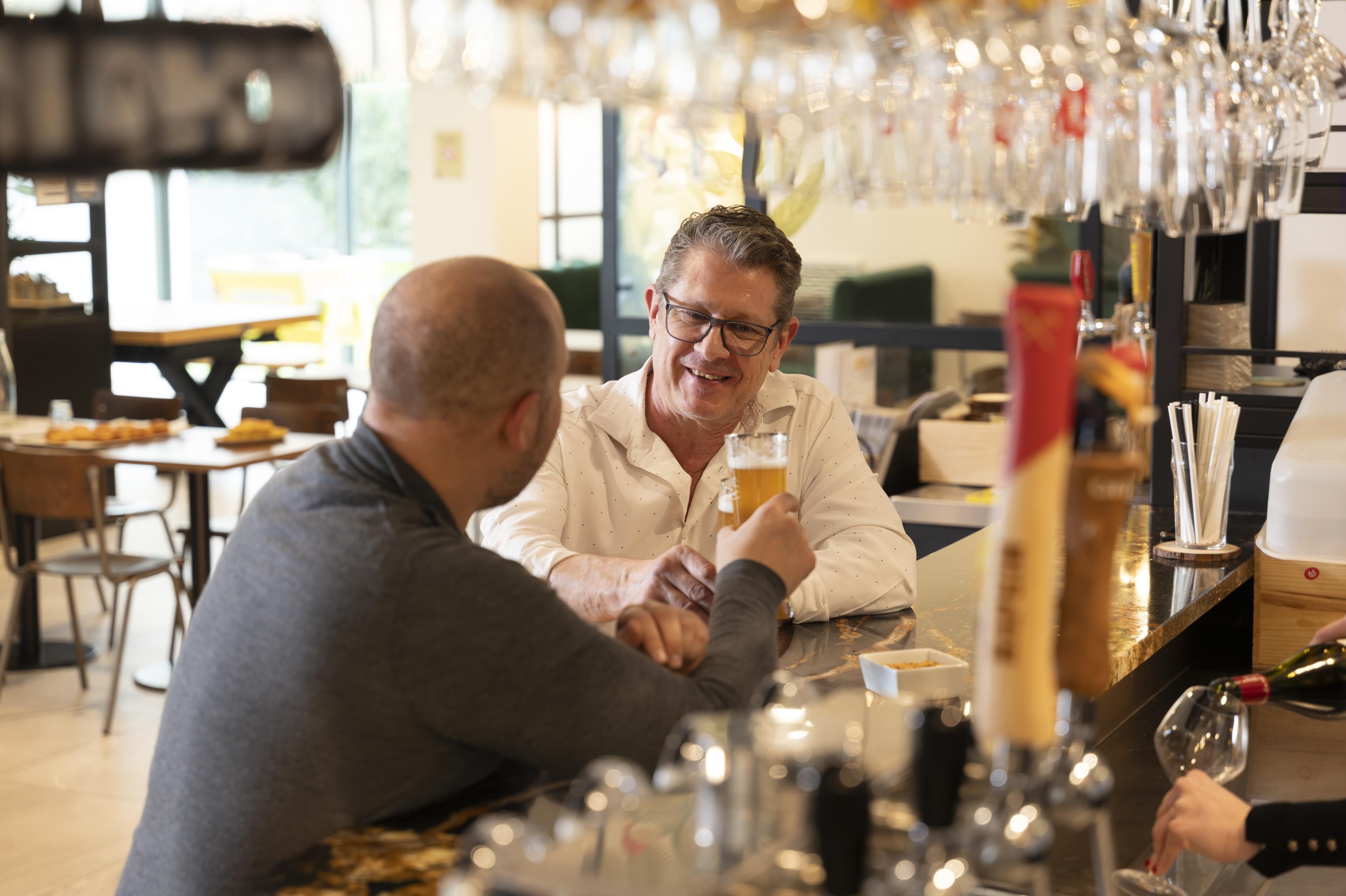 Dos personas sentadas en un bar, con vasos de cerveza, una botella de vino y una barra con vasos y botellas.