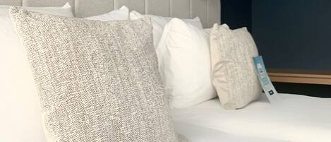 Una cama bien hecha con sábanas blancas y varias almohadas, un cabecero tapizado en gris y una mesita de noche con lámpara.