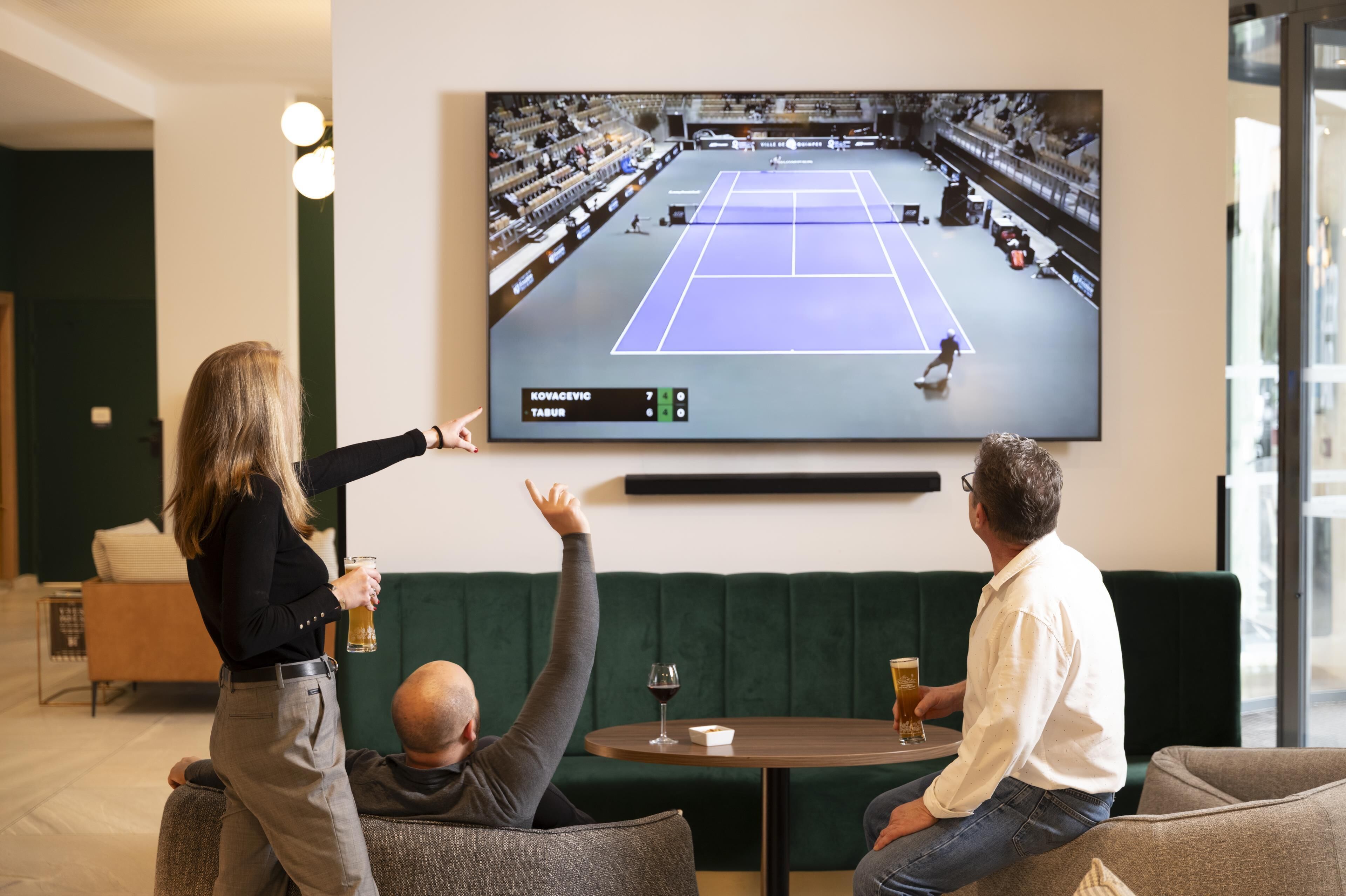 A room with a large flat-screen TV mounted on the wall displaying a tennis match. There are two people seated on a green sofa, one holding a beer and the other a glass of wine.