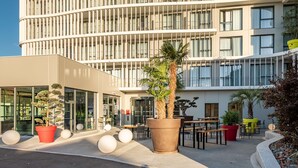 Un edificio moderno con una facciata curva, ampie finestre e aree esterne con piante in vaso e tavoli rotondi.