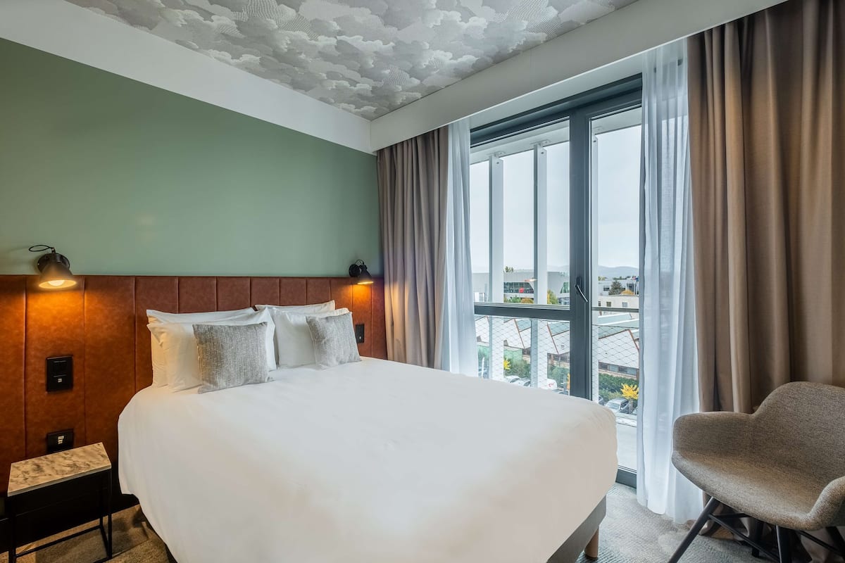 A hotel room with a large bed, a wooden headboard, two wall-mounted lights, a bench, and a view of the city through large windows.