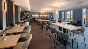 Een moderne eetruimte met meerdere tafels en stoelen, een keuken met planken en planten, en grote ramen die voor natuurlijk licht zorgen.