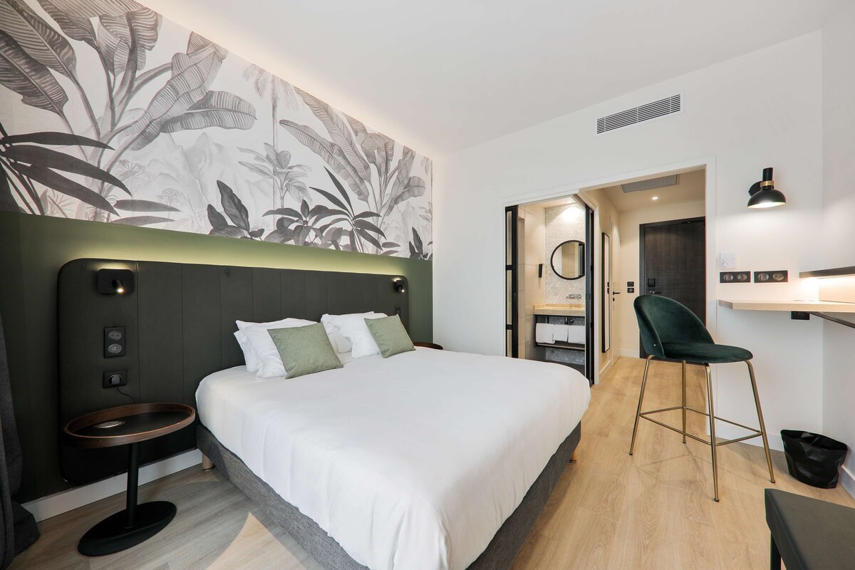Une chambre d’hôtel moderne avec un grand lit, une petite table ronde, un fauteuil vert et un mur orné d’un motif de feuilles.