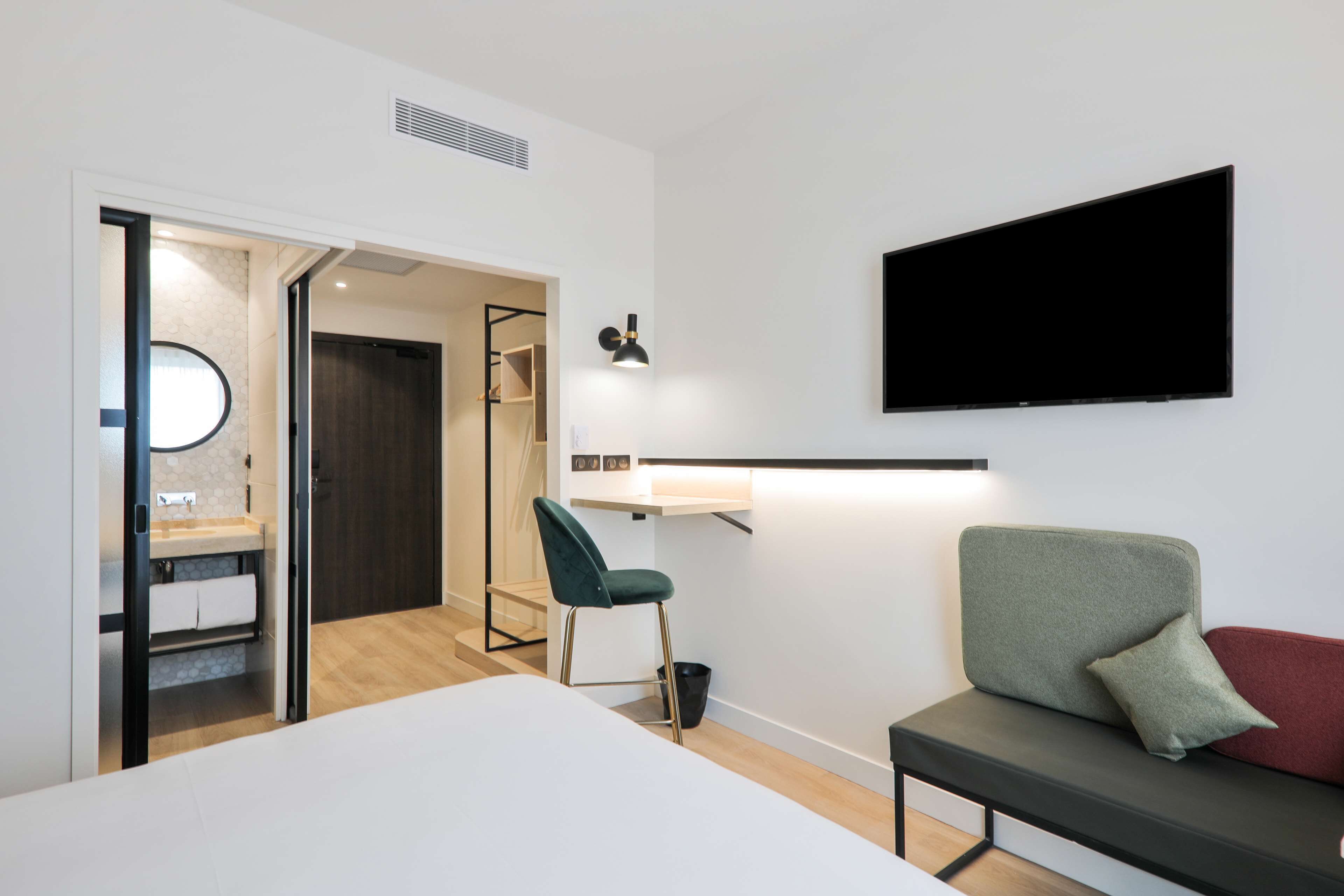 A modern hotel room with a bed, a desk, a chair, a TV, and a bathroom visible through an open door.
