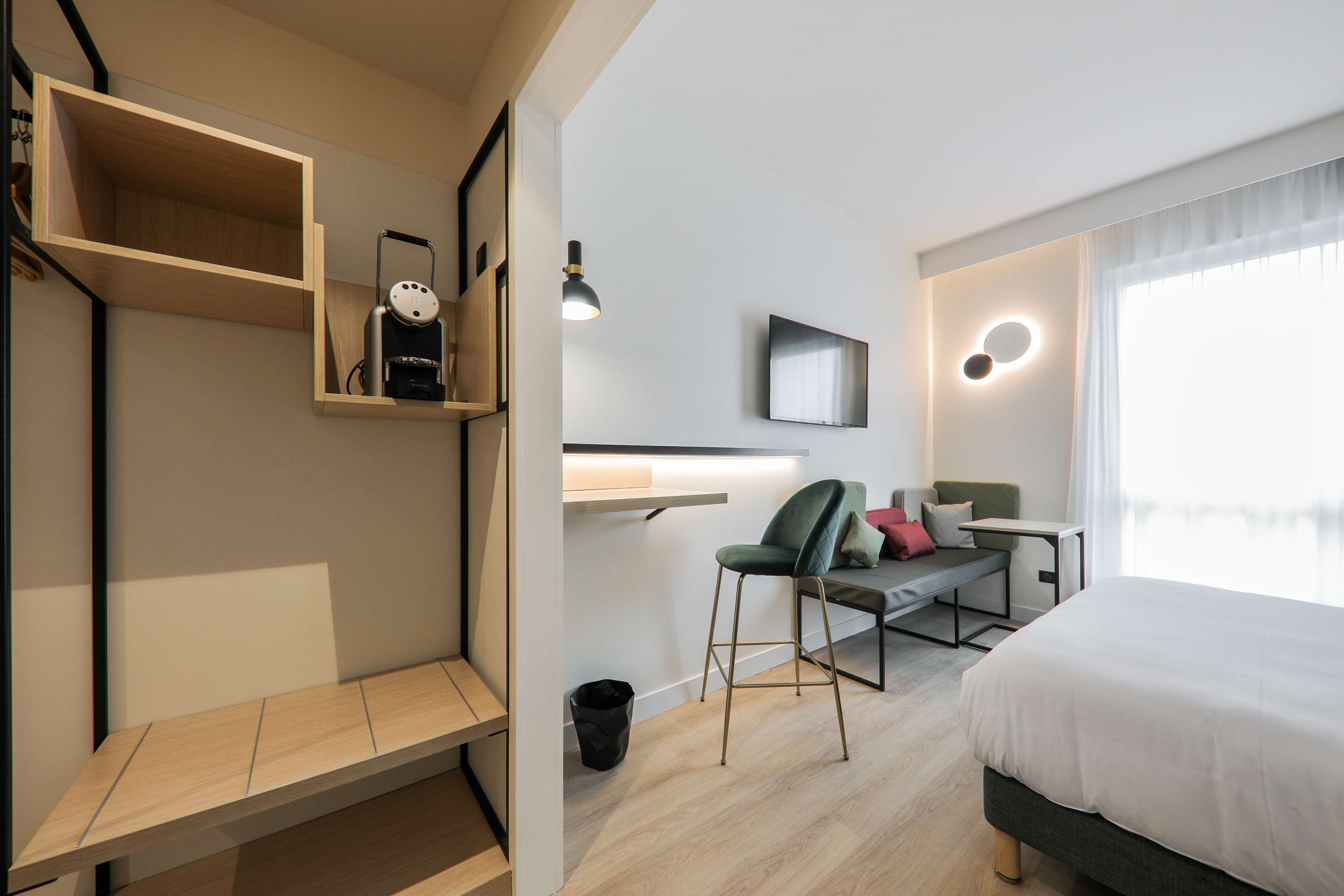 A modern hotel room with a bed, a desk, a chair, a TV, and a shelf with a vacuum cleaner.