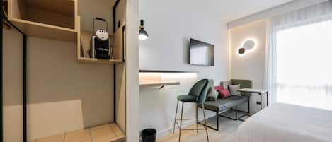 A modern hotel room with a bed, a desk, a chair, a TV, and a shelf with a vacuum cleaner.