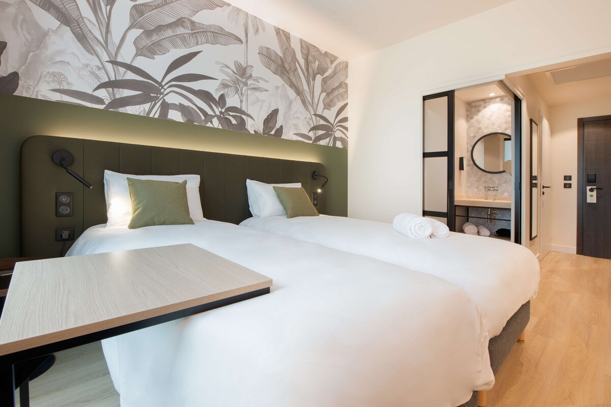 Une chambre d’hôtel avec deux lits, une table de chevet en bois, un miroir et une salle de bain visible à travers une porte ouverte.