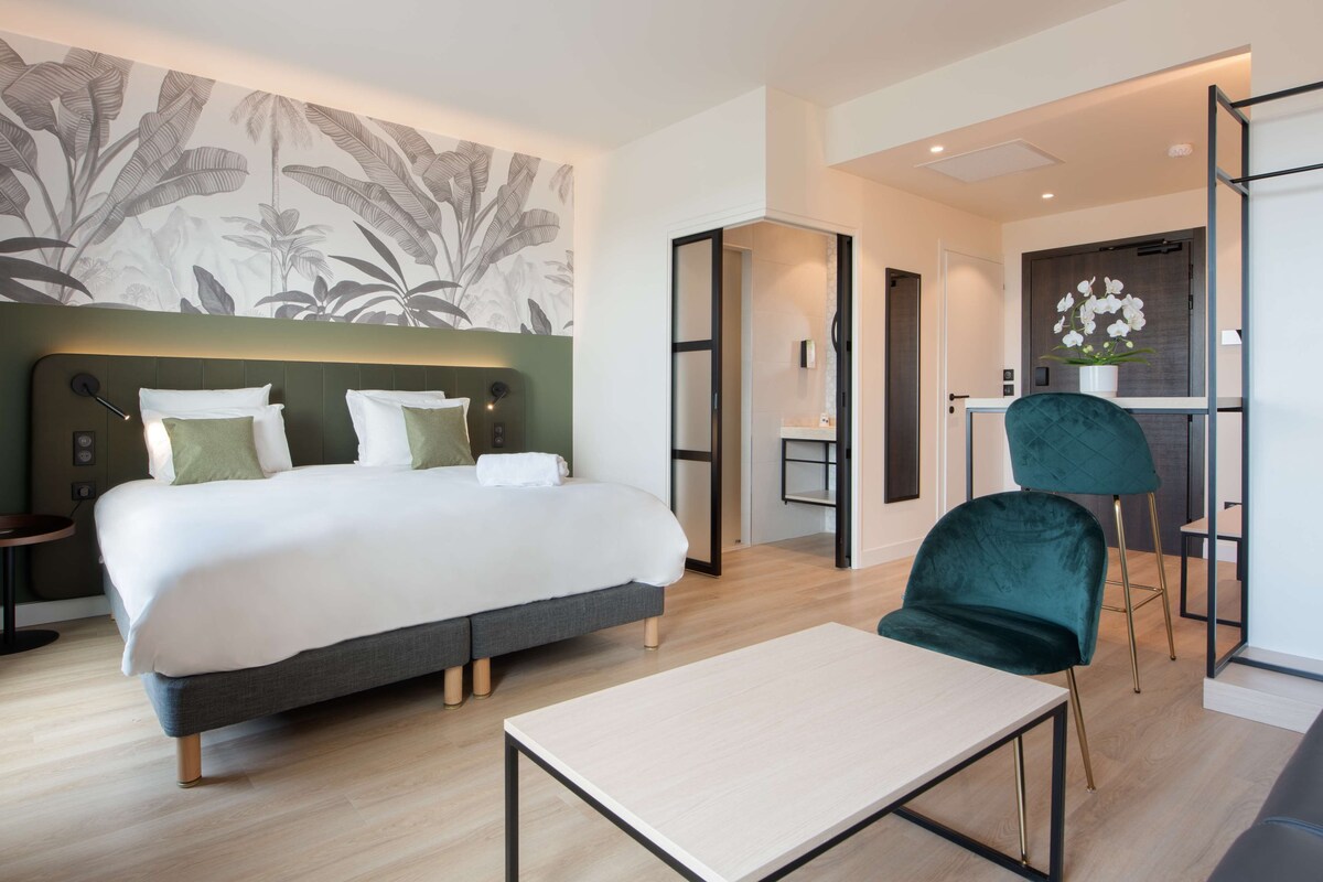 Une chambre d’hôtel moderne avec un grand lit, un bureau et une chaise. Un mur est décoré d’un motif de feuilles et une porte donne accès à une autre pièce.