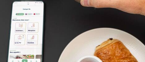 A person holding two drinks with latte art, a smartphone displaying a language app, and a plate with croissants and jam.