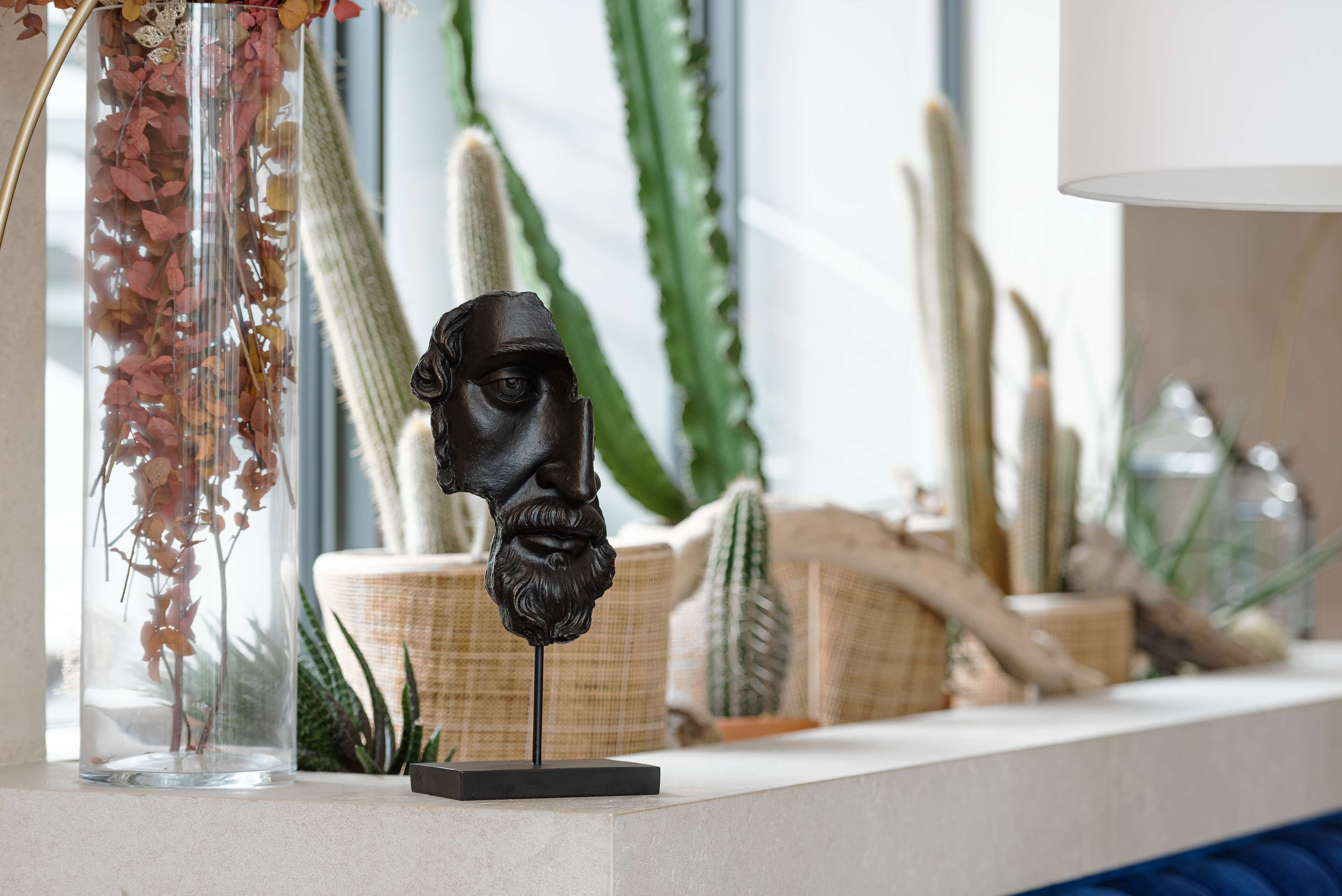 Une sculpture représentant le visage d’un homme, posée sur un socle, entourée de cactus et d’un vase contenant des fleurs séchées.