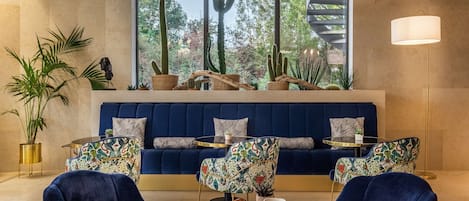 A modern living room with a blue sofa, patterned chairs, a round coffee table, and a large window with a view of cacti and trees.