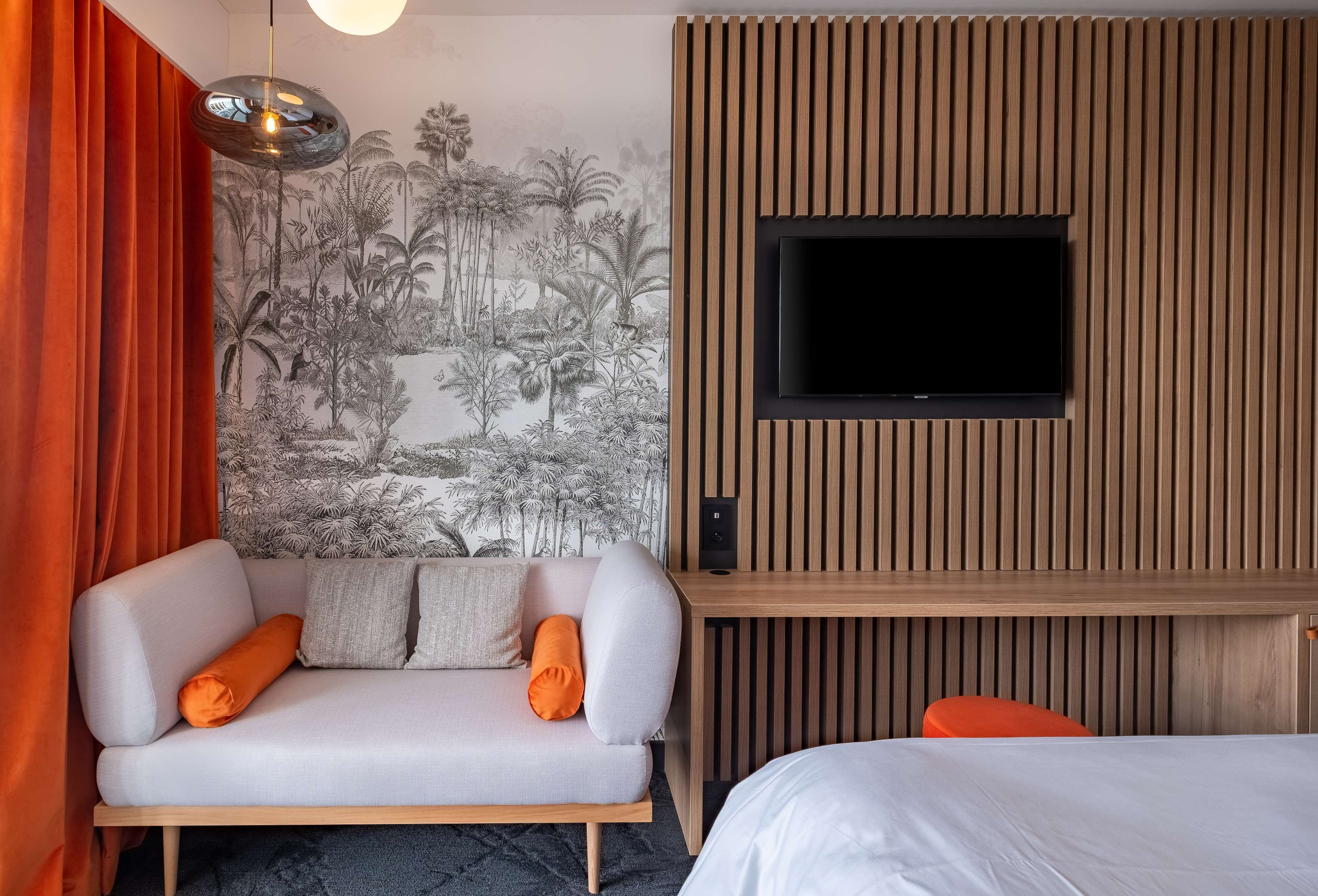 A modern hotel room with a sofa, a bed, a TV mounted on a wooden panel wall, and a mural of tropical scenery.