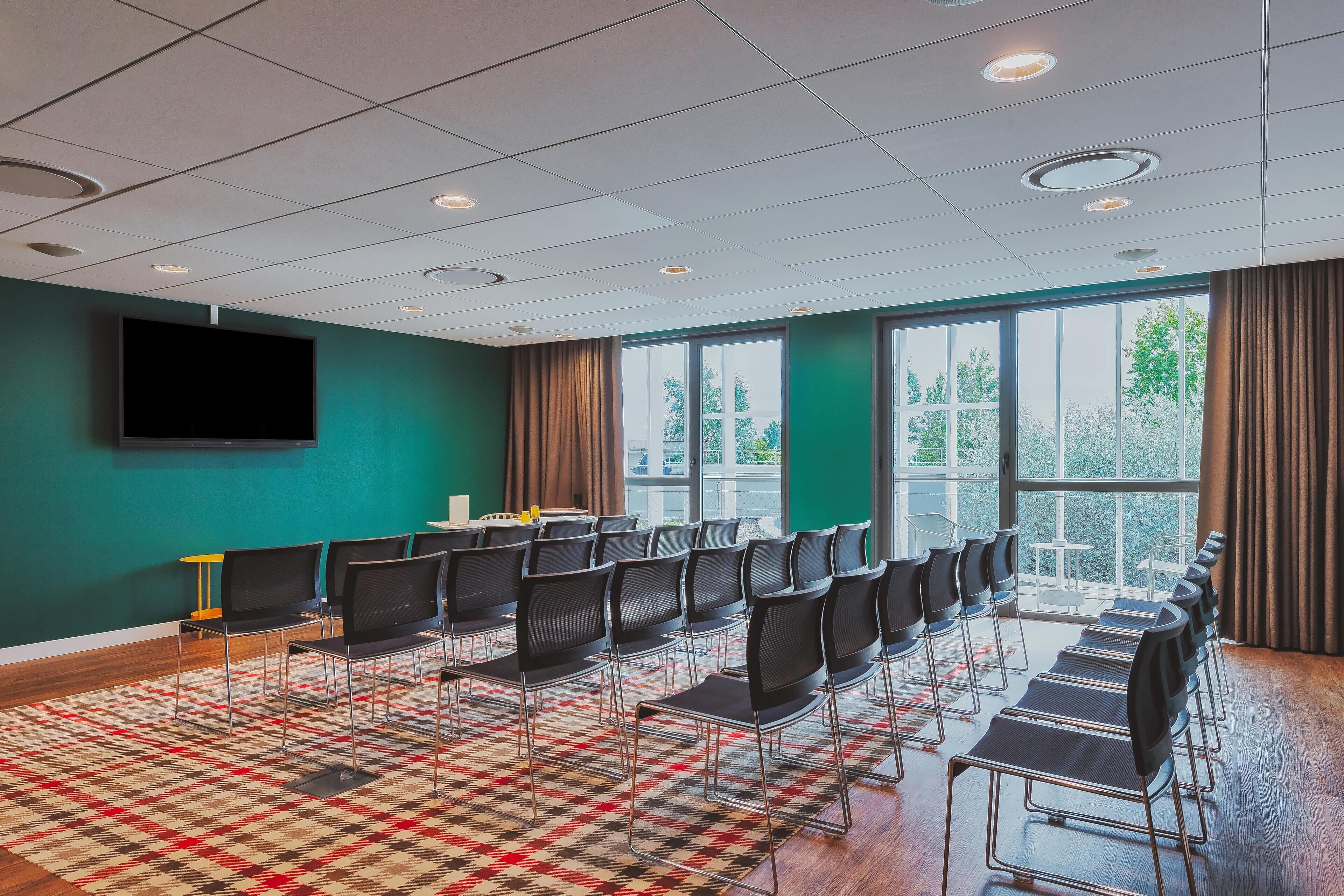 A conference room with rows of chairs, a large window with curtains, a flat-screen TV, and a patterned carpet.