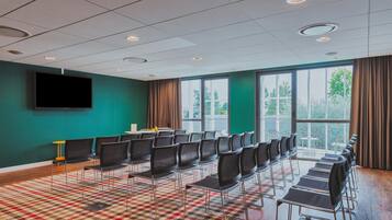 A conference room with rows of chairs, a large window with curtains, a flat-screen TV, and a patterned carpet.