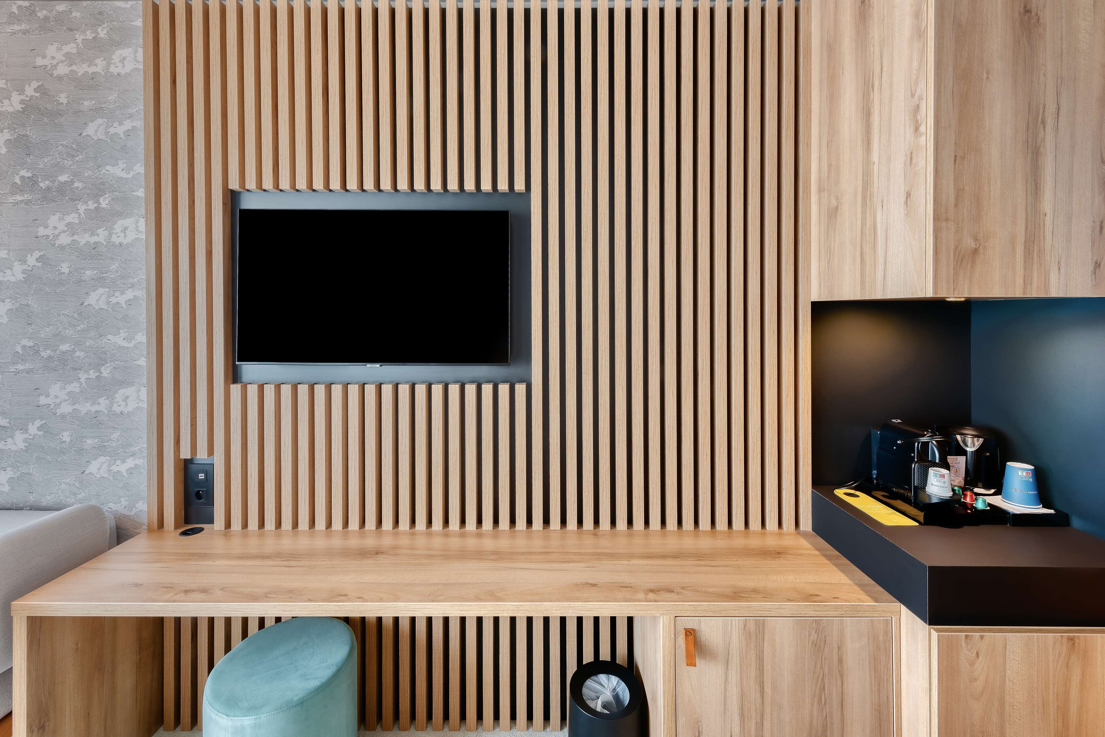 Una habitación moderna con una pared de listones de madera, un televisor de pantalla plana sobre un escritorio de madera, y un pequeño estante con una cafetera y otros objetos.