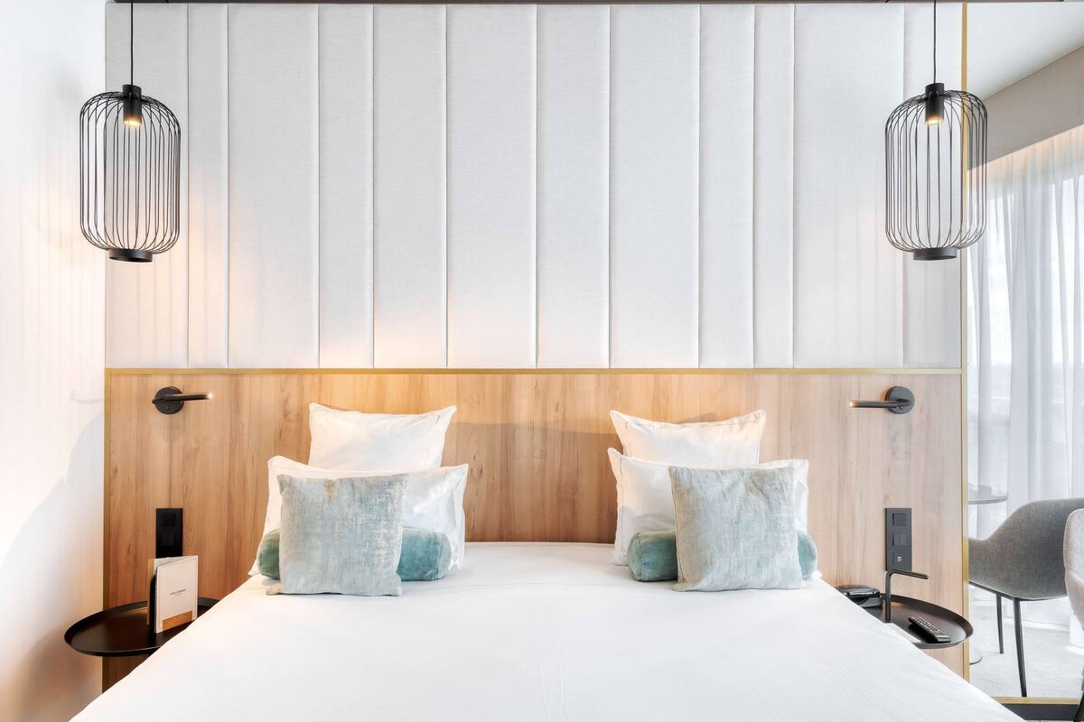 A modern hotel room with a large bed, two bedside tables, a wooden headboard, and two pendant lights.