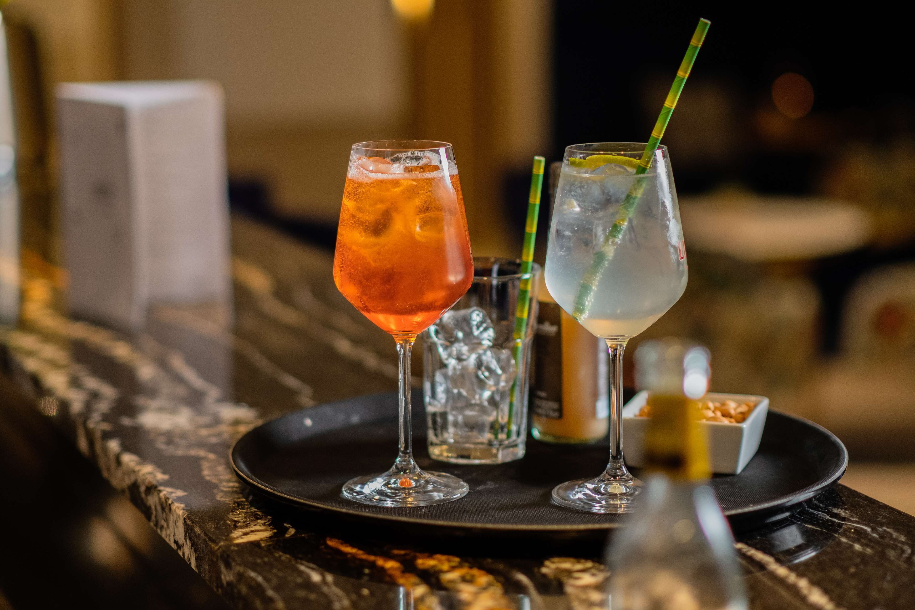 Deux verres de cocktails sur le comptoir en marbre d’un bar, accompagnés d’un petit bol de noix et d’une bouteille de spiritueux.