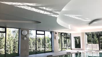A modern indoor swimming pool with large windows, a curved ceiling, and a clear view of the surrounding greenery.