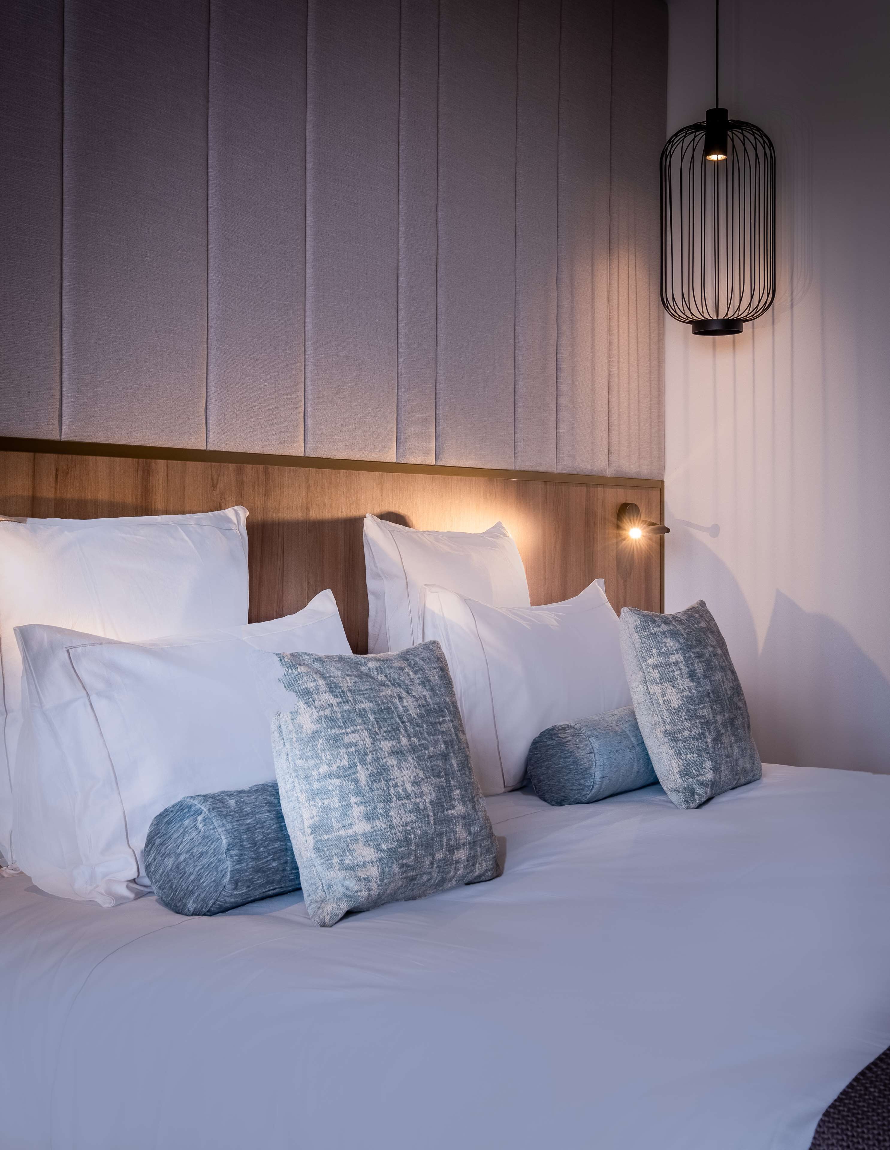 A neatly made bed with white and grey pillows, a wooden headboard, and a hanging light fixture.