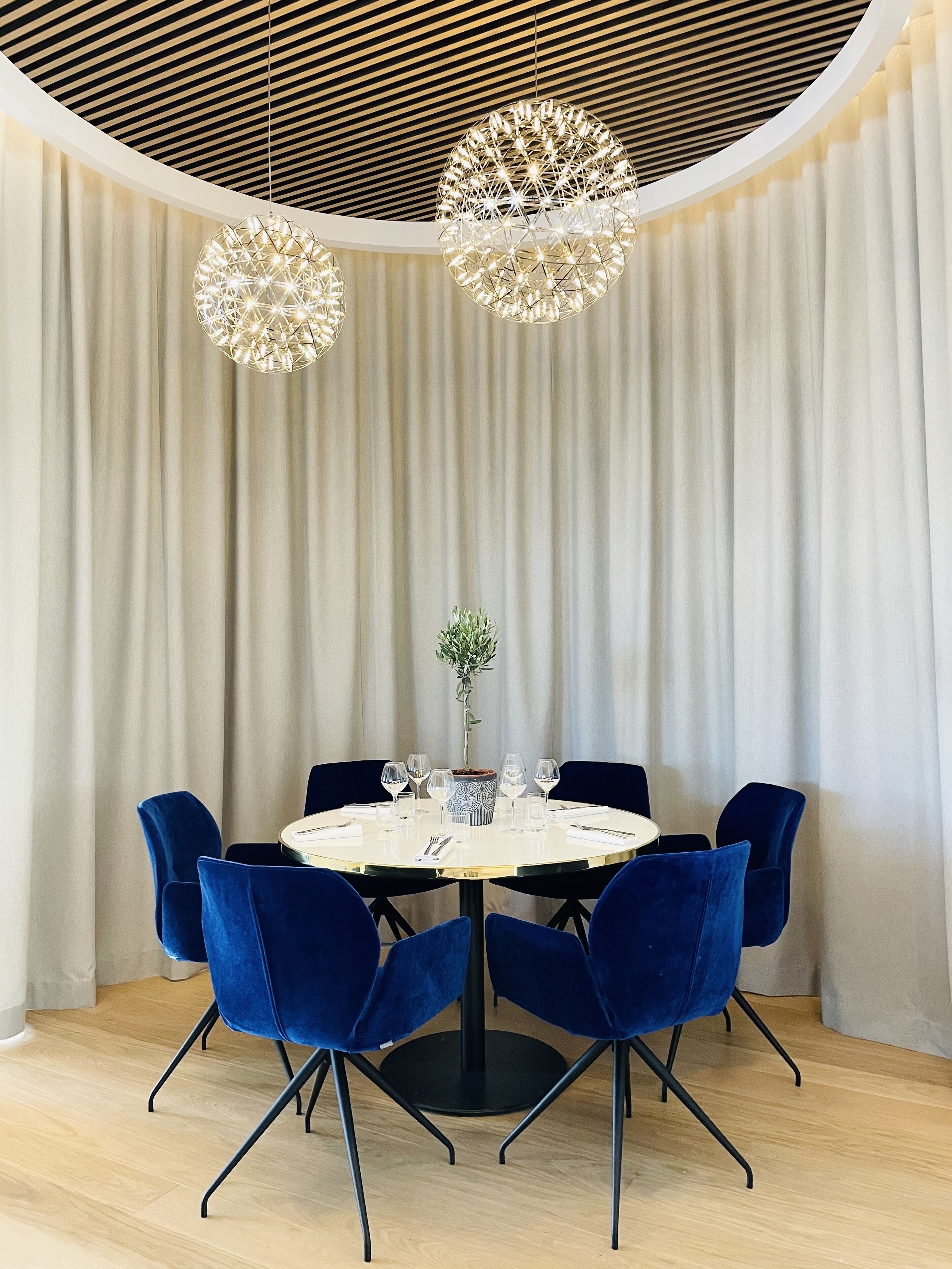 Een ronde eettafel met zes blauwe stoelen, een potplant en wijnglazen op een witte tafelkleed.