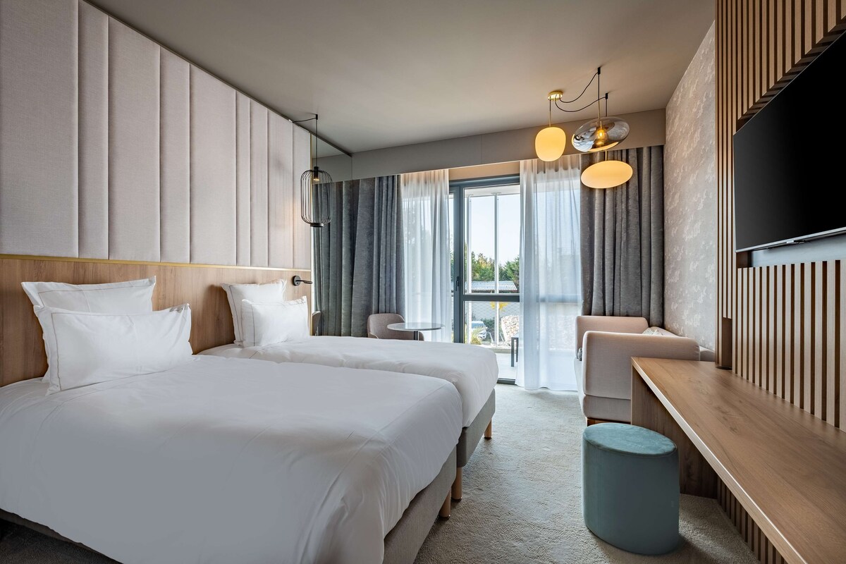 Une chambre d’hôtel moderne, équipée de deux lits, d’un banc en bois, d’une table ronde et d’un téléviseur fixé au mur.